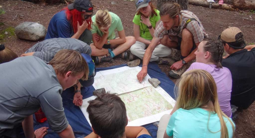 A group of students sit around a map spread out on the ground. One person, who appears to be an instructor, points at the map.